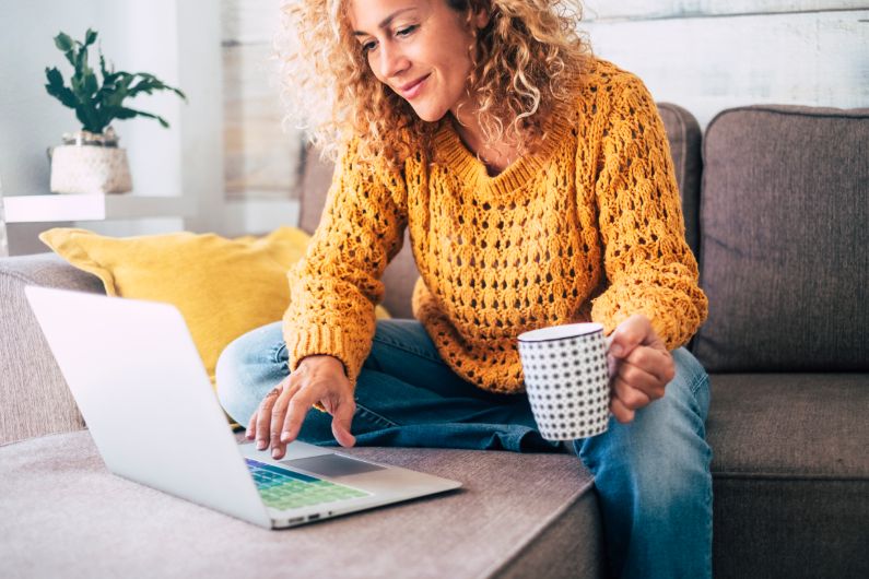 a woman in a yellow sweater smiles at a laptop on the coffee table while holding a coffee mug