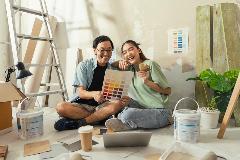 A husband and wife sits on the floor of their home looking at paint colors
